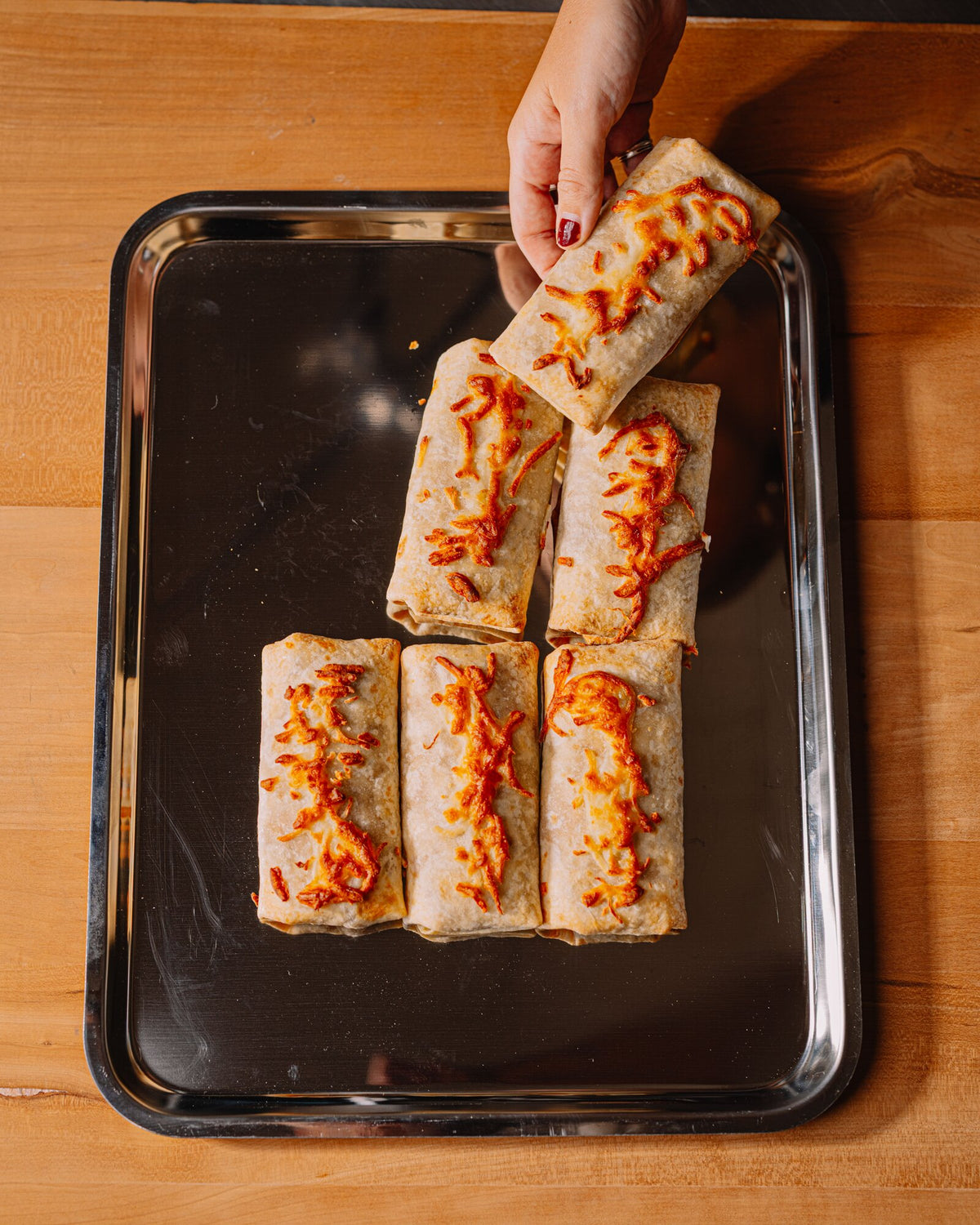 Six Carne con Chile burritos on a baking tray, with a hand lifting one burrito